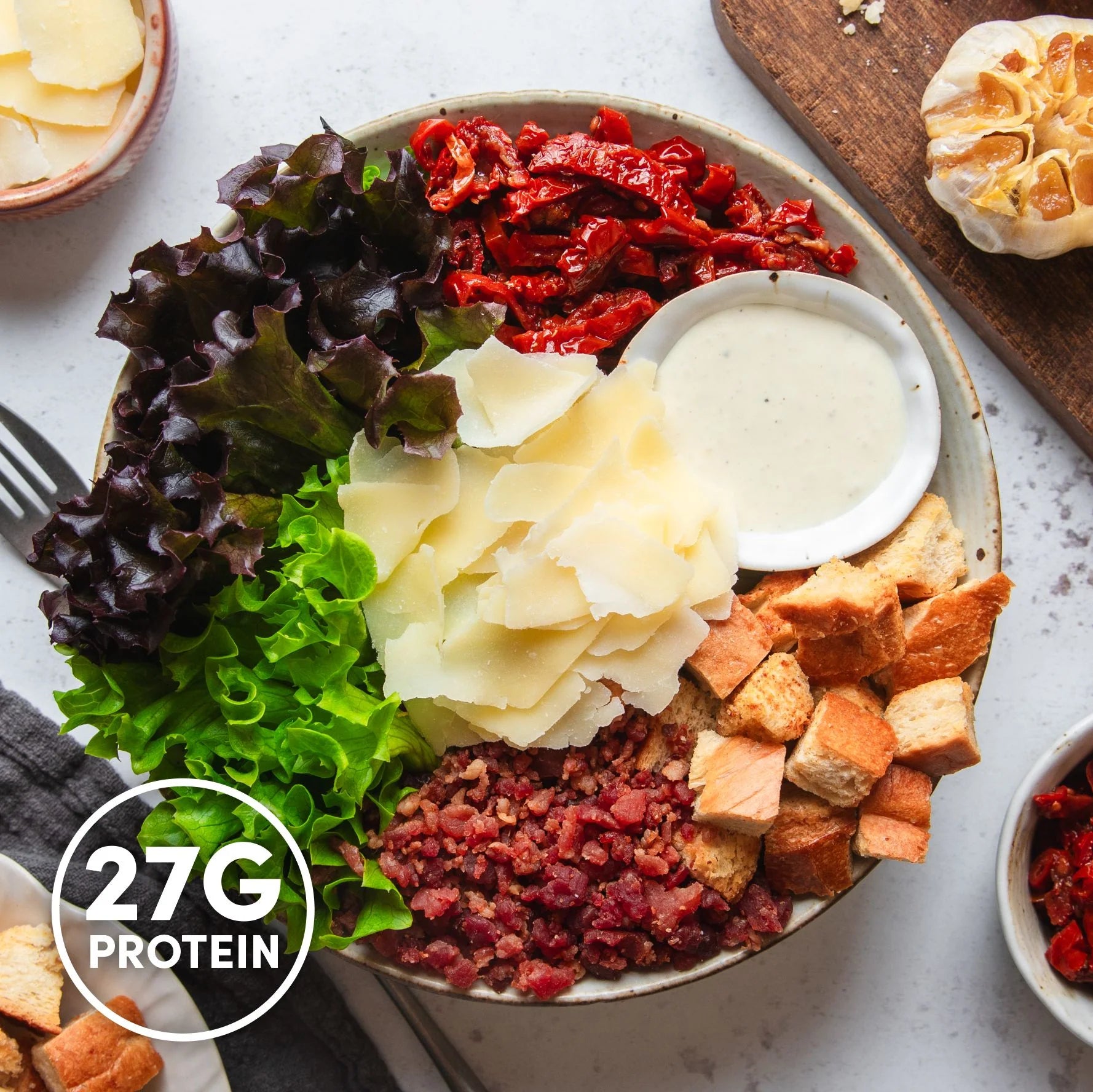 Close-up of a high-protein Caesar-style salad bowl with crispy bacon bits, shaved parmesan, fresh lettuce, croutons, sun-dried tomatoes and dressing, labelled 27 grams of protein.